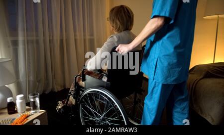 Infermiera che porta la vecchia sedia a rotelle depressa alla finestra che allatta la solitudine domestica Foto Stock