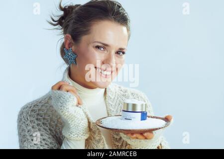 Ritratto di felice casalinga elegante in rullo pullover collo e cardigan piastra di tenuta con il viso creme tra neve contro inverno sfondo azzurro. Foto Stock