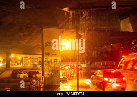 L'uomo senza tetto cucina sotto un viadotto in una notte piovana nel centro di Rio de Janeiro - cabina di polizia abbandonata in background. Foto Stock