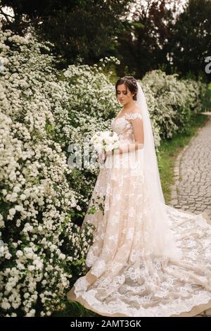 Sposa di charme immerso in uno splendido abito da sposa tenere bouquet e stare di fronte ad alberi in fiore Foto Stock