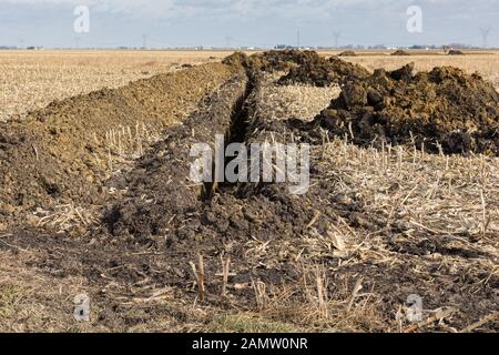 Trincea scavata nel campo di fattoria con cumuli di terra. L'acqua della tubazione di drenaggio, campo di installazione di piastrelle Foto Stock