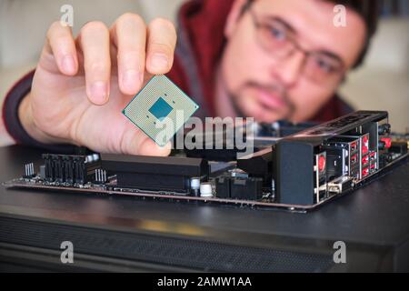 Uomo che esamina un chip di computer tenuto tra le dita, sopra una scheda madre. Foto Stock