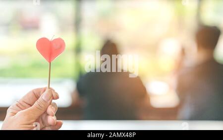 Mano di donna tenere una candela a forma di cuore rosso con un'immagine sfocata di coppia sullo sfondo. San Valentino e concetto di amore. Foto Stock