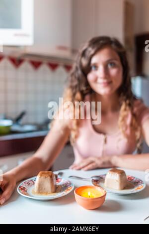 Dolce panna cotta cibo dessert con bokeh di giovane donna felice sorridente seduto al tavolo da cucina guardando la macchina fotografica al chiuso in Italia data romantica Foto Stock