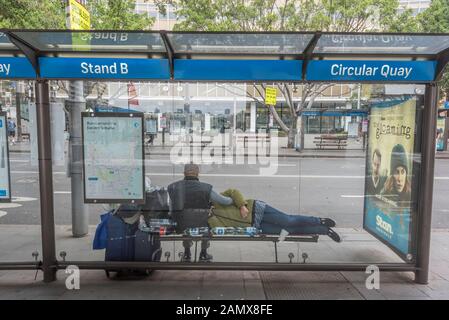 Una donna di mezza età si trova con la sua testa in un uomo di giro in attesa con i loro bagagli in corrispondenza di una fermata di autobus a Circular Quay nel centro di Sydney, Australia Foto Stock