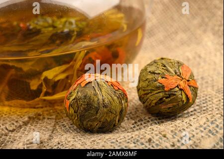 Tè dei fiori preparato in una teiera di vetro e palle da tè dei fiori su uno sfondo di tessuto ruvido homespun. Primo piano Foto Stock