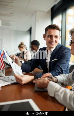 Giovane imprenditore sorridente puntando al documento e di discuterne con il suo partner al tavolo durante la conferenza Foto Stock