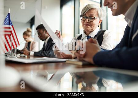 Sorridente imprenditrice senior in occhiali seduti a tavola la lettura di relazione e a parlare con il suo collega a una conferenza Foto Stock