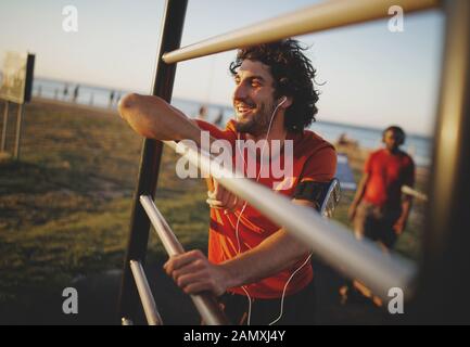 Ritratto di un sorridente giovane maschio atletico godendo la musica dal suo auricolari riposo dopo gli esercizi in palestra all'aperto Foto Stock