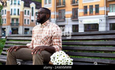 Giovane uomo in attesa di donna, seduto su panchina parco con fiori, data cieca Foto Stock