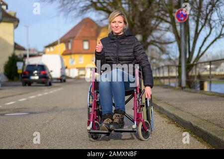 Sorridente donna disabili con sedia a rotelle in una strada urbana dando un pollice su per dimostrare il suo atteggiamento positivo Foto Stock
