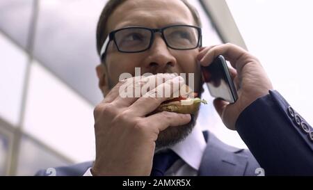 Nutrizione non sana, busy busy businessman parlare al telefono e mangiare hamburger pranzo Foto Stock