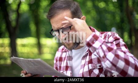 Uomo che invecchia in occhiali cercando di leggere giornale nel parco, lungimiranza, miopia Foto Stock