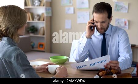 Marito occupato nell'abito che discute le statistiche dell'azienda sul cellulare, ignorando la moglie Foto Stock