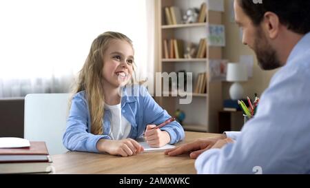 Sorridendo piccola figlia seduto al tavolo e facendo i compiti con il padre, la cura Foto Stock