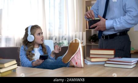 Il padre occupato che dà i soldi della tasca alla figlia capricciosa piccola con le cuffie Foto Stock