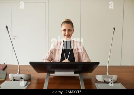 Ritratto di giovane imprenditrice di successo di sedere a tavola e sorridente in telecamera lei parlando alla conferenza di business Foto Stock