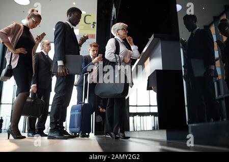 Donna senior con i bagagli in piedi vicino al ricevimento e registrazione con altre persone in piedi in una coda in aeroporto Foto Stock
