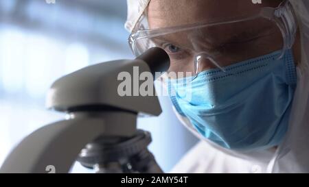 Assistente di laboratorio maschile in maschera protettiva e occhiali utilizzando il microscopio per la ricerca Foto Stock