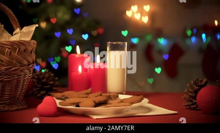 Bicchiere di latte e biscotti allo zenzero sul tavolo, albero di Natale frizzante con luci Foto Stock