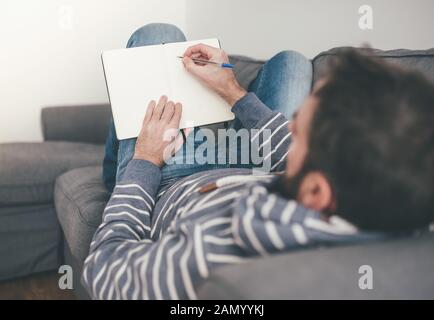 uomo che si rilassa sul divano mentre prende appunti o scrive in diario o notebook Foto Stock