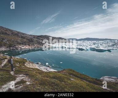 Vista verso Icefjord a Ilulissat. Giovane viaggiatore turistico in piedi sul lungomare. Iceberg del ghiacciaio Kangia in Groenlandia nuoto con il cielo blu Foto Stock