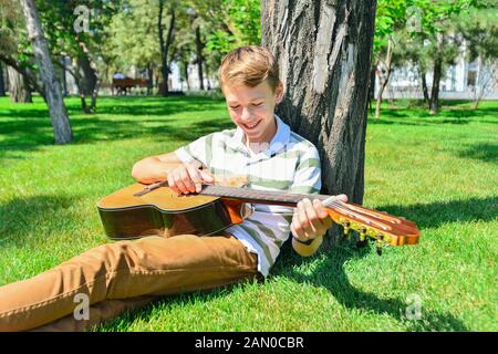 Un ragazzo con una chitarra sotto un albero, un adolescente suona e canta nel parco. Foto Stock