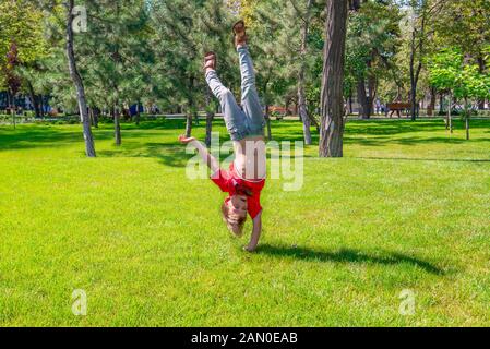 Un ragazzo si erge su un braccio a testa in giù in un parco sul prato verde. Foto Stock