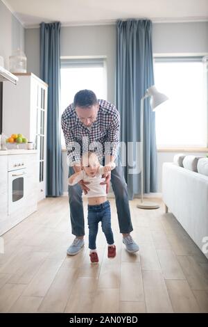 Padre nella camicia a scacchi aiutare la sua bimba a camminare Foto Stock