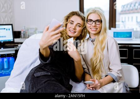 Bella Donna sorridente in grigio morbido accappatoio e il suo medico personale cosmetologo, tenendo creme, sorridente e divertirsi, rendendo selfie foto in Foto Stock