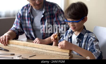 Attento ragazzino martellante chiodo in legno Plank, padre che sostiene il suo ragazzo Foto Stock