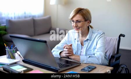 Donna anziana positiva in sedia a rotelle che lavora su laptop, IT freelancer, online Foto Stock