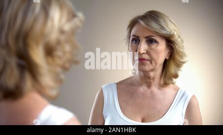 Femmina pensionato guardando nello specchio e pensando alla chirurgia plastica, riflessione Foto Stock