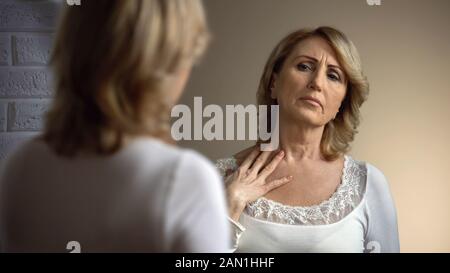 Donna senior infelice che guarda nello specchio e tocca la zona del decollito, le rughe Foto Stock