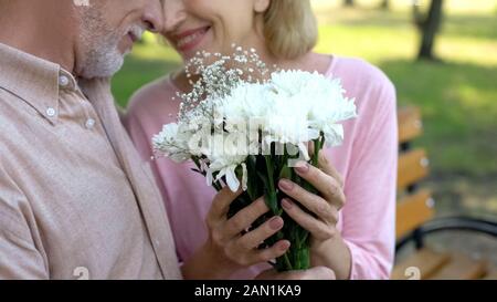 Felice Coppia matura nuzzling, tenendo i fiori bianchi bouquet, seduto in una panchina nel parco Foto Stock