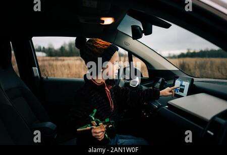 il ragazzo sedette sul sedile del conducente di un'auto elettrica utilizzando il navigatore satellitare Foto Stock