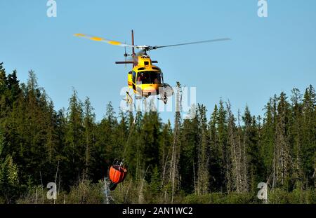 Un elicottero con un secchio d'acqua appena riempito che si solleva da un lago remoto nella zona rurale dell'Alberta e vola per combattere un vicino incendio boschivo Foto Stock