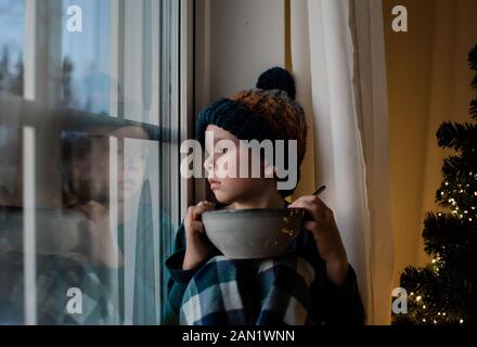 il ragazzo sedette su un davanzale con una ciotola di cereali che guarda all'esterno Foto Stock