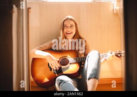 Giovane allegra donna che tiene la chitarra mentre si siede sul pavimento del balcone Foto Stock
