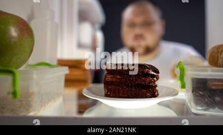 Uomo Oversize tenendo pezzo di torta dal frigo durante la notte, il rischio di diabete, calorie Foto Stock