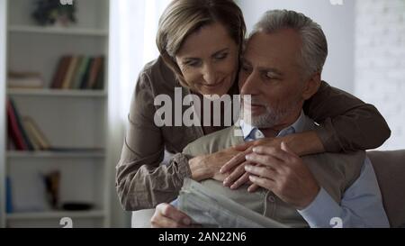 Anziani dando femmina abbraccio per marito seduto sul divano e quotidiano di lettura Foto Stock