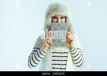 felice trendy 40 anni donna in bianco maglione a righe, sciarpa e orecchie flap cappello nascosto dietro notebook isolato su sfondo inverno azzurro. Foto Stock