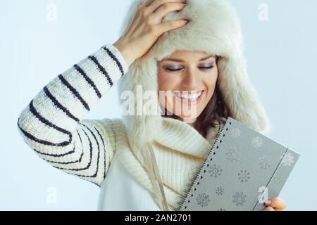 allegra femmina moderna in maglione a righe bianche, sciarpa e cappellino con taccuino su sfondo blu chiaro invernale. Foto Stock