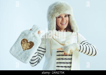 Ritratto di felice donna moderna in maglione a righe bianche, sciarpa e cappellino auricolare che punta a cute calda acqua calda riempimento caldo isolato su luce invernale bl Foto Stock