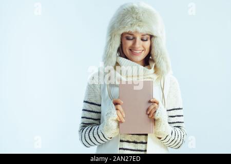 giovane donna sorridente con maglione a righe bianche, sciarpa e cappello con alette auricolari che guarda un libro isolato su sfondo azzurro invernale. Foto Stock