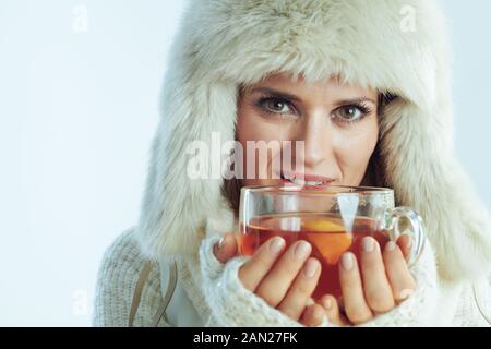 Ritratto di donna moderna in maglione a righe bianche, sciarpa e lembo auricolare cappello bere tazza di tè caldo con limone e miele contro inverno azzurro schiena Foto Stock