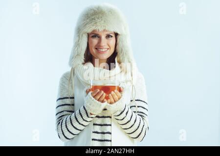 Ritratto di donna sorridente alla moda in maglione a righe bianche, sciarpa e lembo auricolare cappello che tiene una tazza di tè caldo con limone e miele isolato su ligh inverno Foto Stock