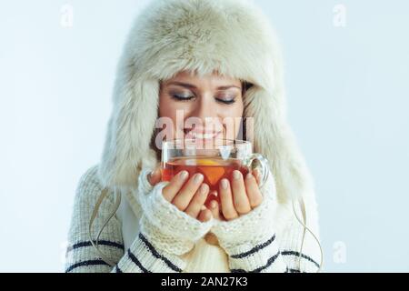 felice giovane di 40 anni donna in maglione a righe bianche, sciarpa e cappello auricolare godendo tazza di tè caldo con limone e miele in inverno blu chiaro bac Foto Stock