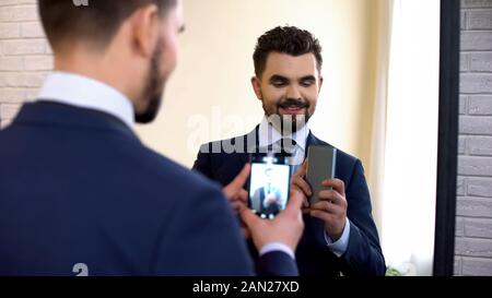 Allegro sorridente nel maschio convenzionale prendendo selfie nella riflessione speculare a casa Foto Stock