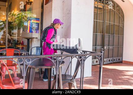 Una femmina di mezza età tende la gamba mettendola su una ringhiera in ferro battuto sul patio di una caffetteria prima di allenarsi. Foto Stock